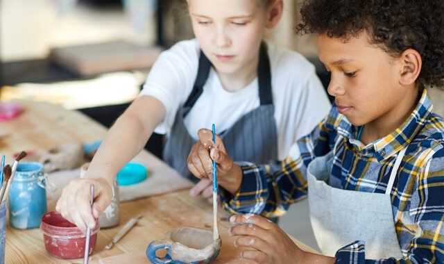 Two youthful classmates sitting by table and painting self-made clay items at lesson